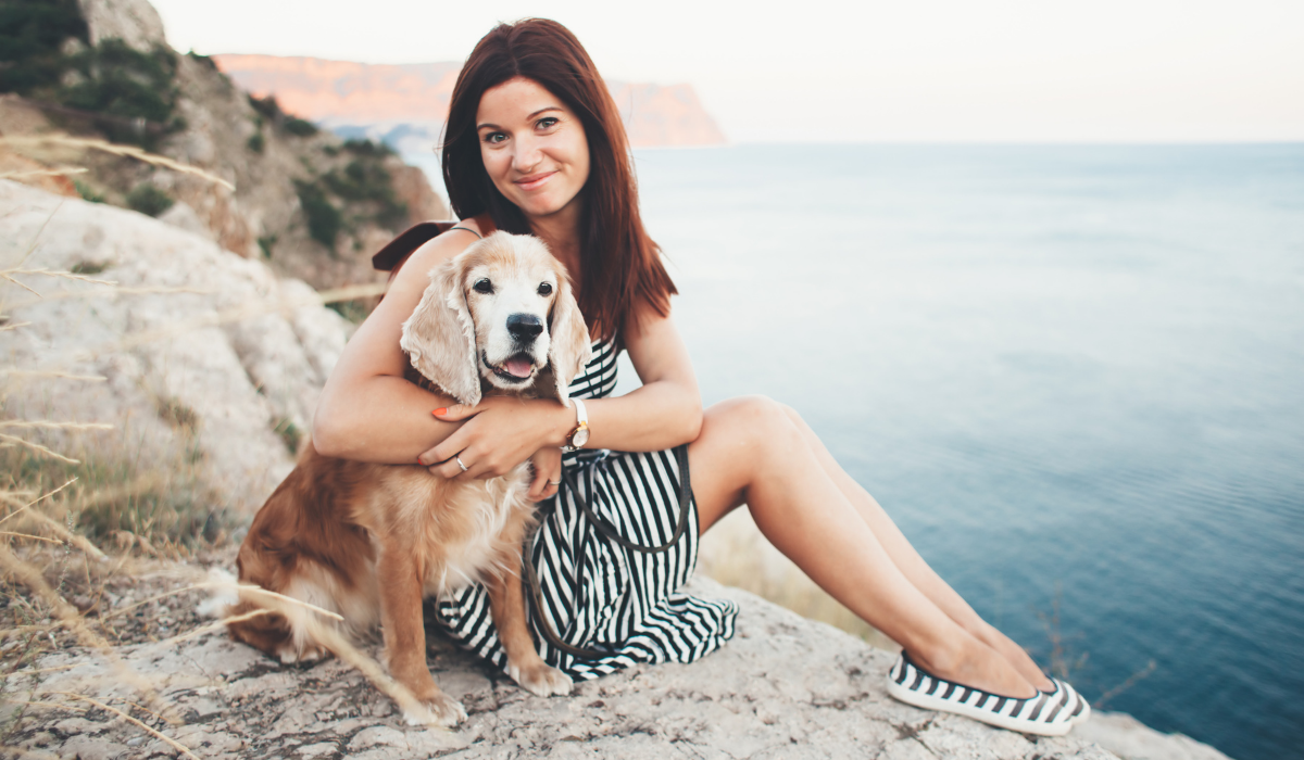 A smiling woman sitting by the ocean holding her spaniel dog wearing a blue and white Evil Eye protection charm.
