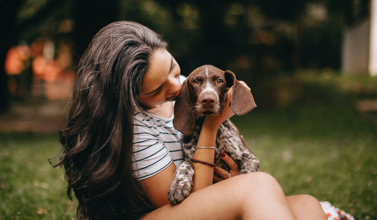 A woman sitting on a park lawn lovingly holding her brown German Shorthaired Pointer puppy wearing an Evil Eye dog collar for protection.
